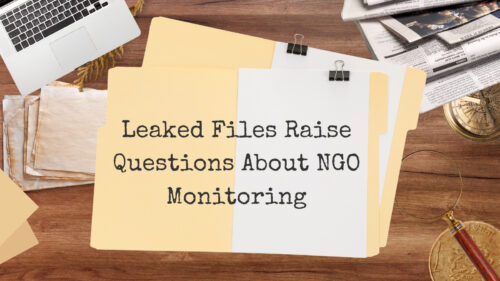 Desk with yellow folders and a white sheet bearing the headline 'Leaked Files Raise Questions About NGO Monitoring' on a wooden surface with a laptop, papers, and a magnifying glass nearby.