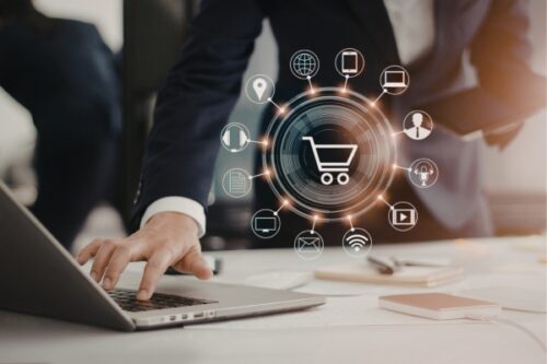 Person typing on a laptop with a glowing shopping cart hub and surrounding digital icons representing online commerce networks.