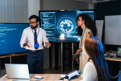 Three coworkers in a tech briefing, with a man in a blue lanyard presenting beside a large monitor showing code and a globe graphic.