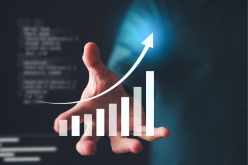 Hand reaching under a rising bar chart with an upward arrow, symbolizing business growth and progress.