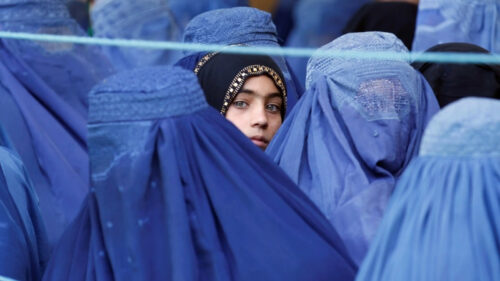 Central woman in a black headscarf with gold trim peeks out from a crowd of women wearing blue robes and veils.